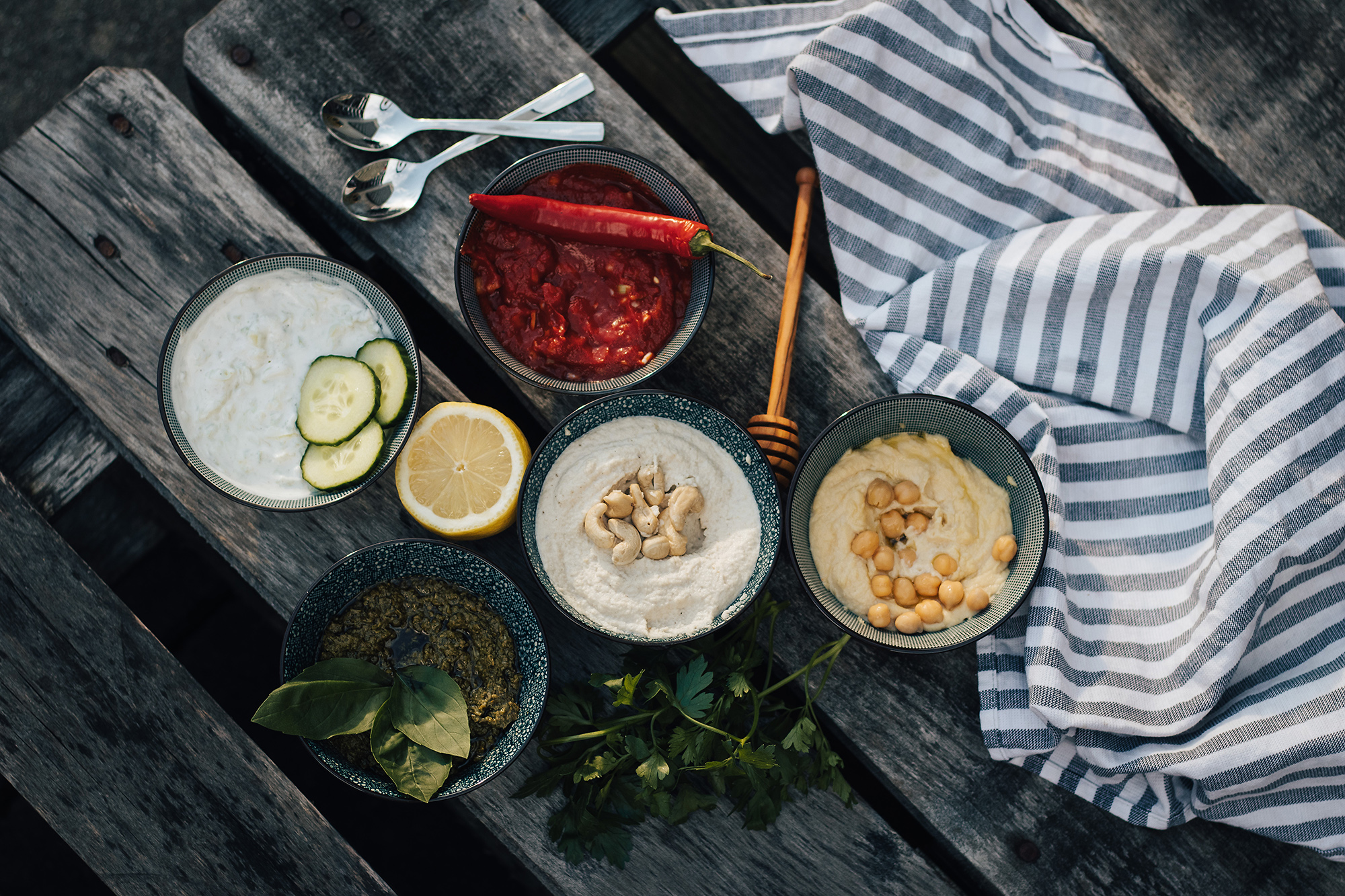 Dip-Rezepte sind vielfältig! Fünf Dips (BBQ, Tzatziki, Hummus, Pesto) auf Holztisch mit Zitrone.